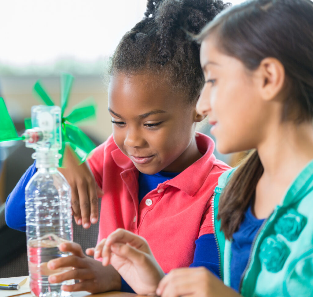 African American and Hispanic elementary age little girls are building a windmill from a water bottle during an after school science program in a public elementary school library. Students are learning science, technology, engineering, and math while working on science project together.