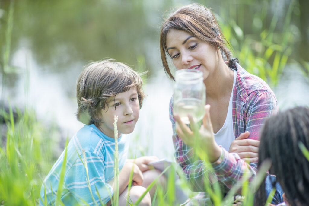 A female teacher holds up a jar of pod water as she teaches two students about the ecosystem around them during an outdoor science class.  They are each dressed casually and the students are looking carefully as the teacher describes what they are seeing.