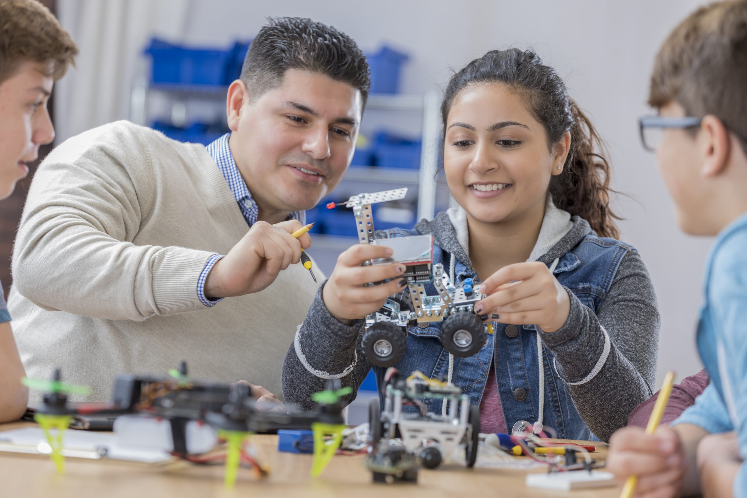 Confident mid adult Hispanic teacher helps a female student with a robotics assignment. The student is holding a robotic vehicle. Robot parts are on the table.